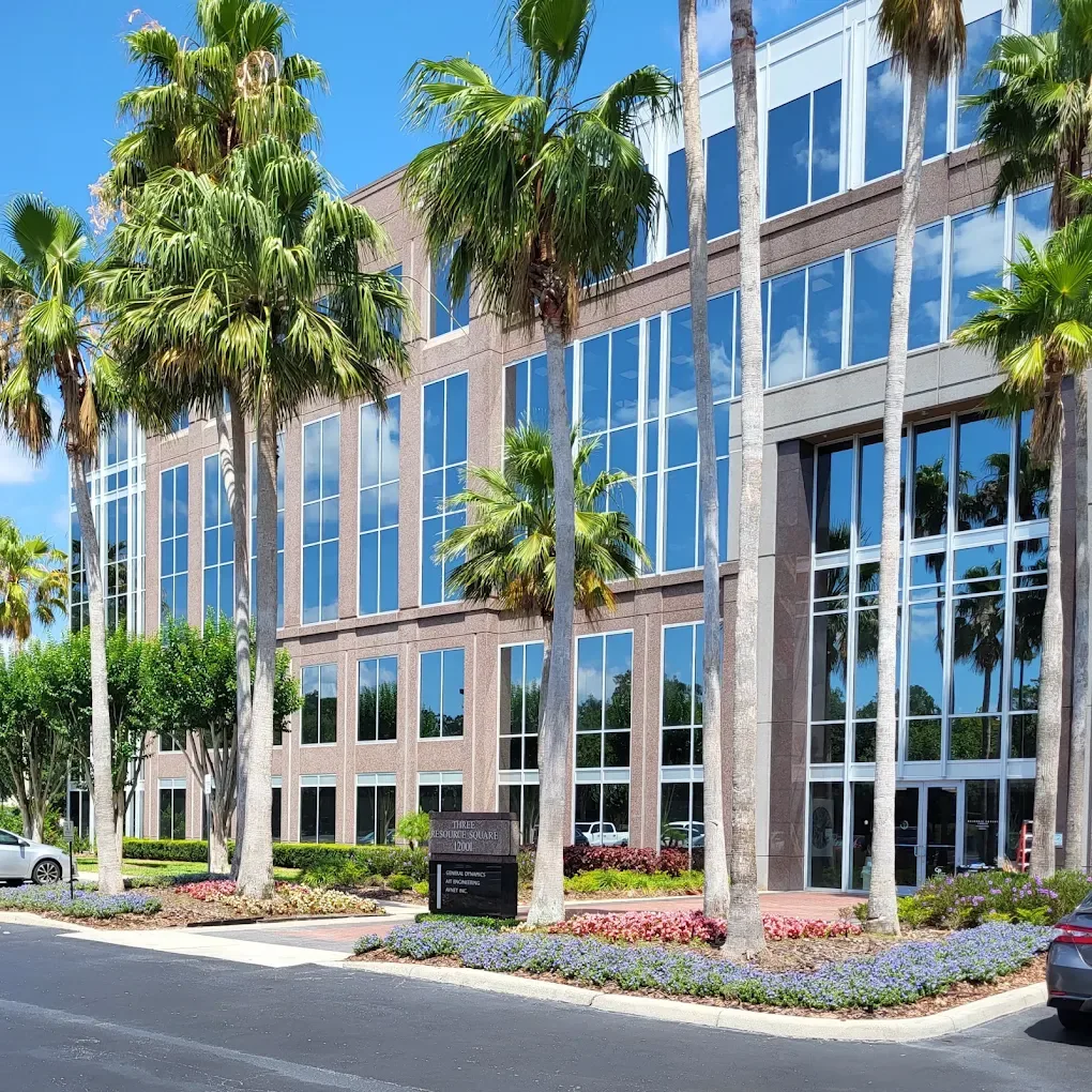Modern brick and glass office building with tall palm trees and colorful landscaping in front.