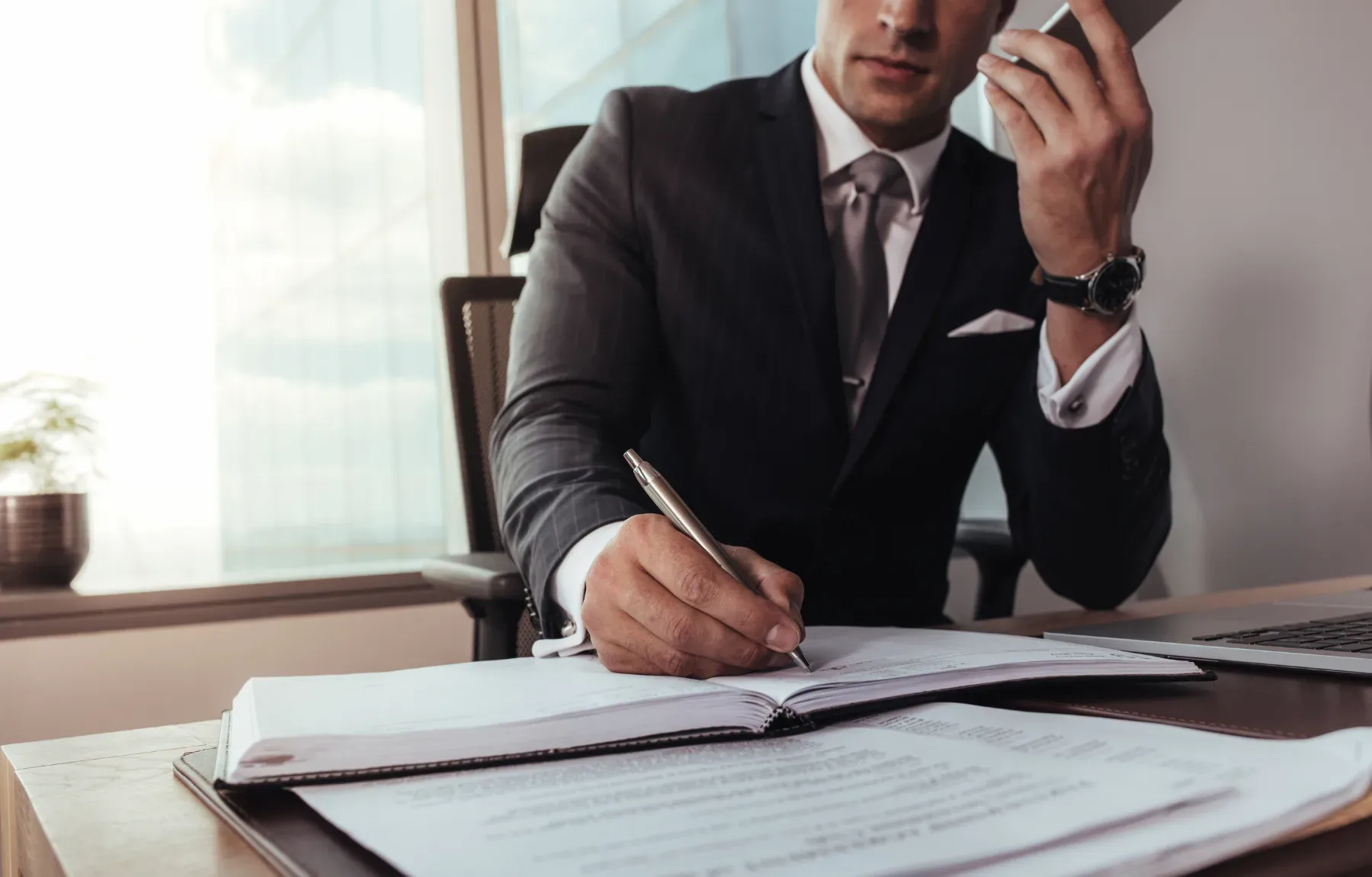 Professional businessman in formal attire writing in a notebook while on a phone call in a modern office setting, representin